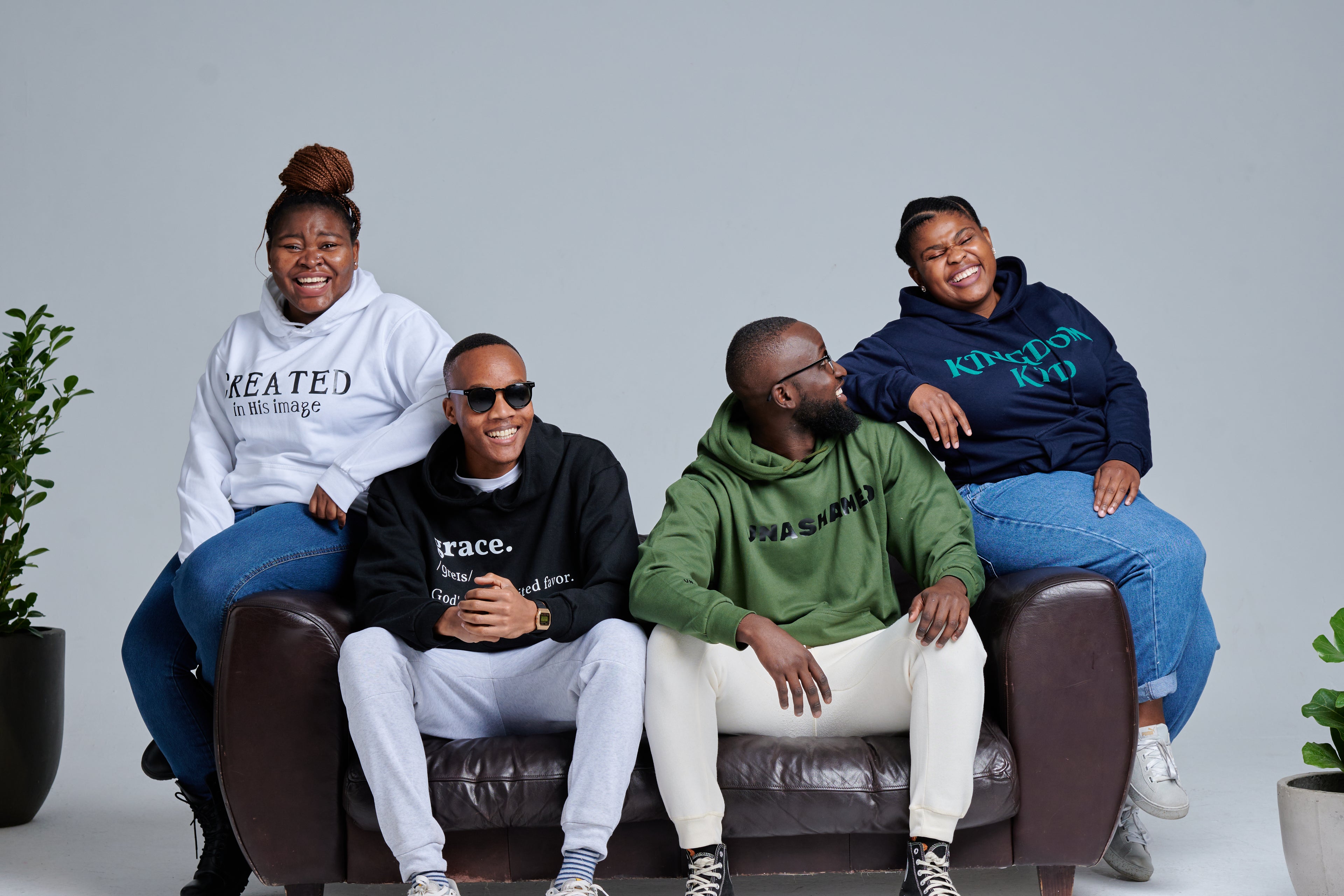 Four people sitting on a couch wearing hoodies with text, against a plain background.